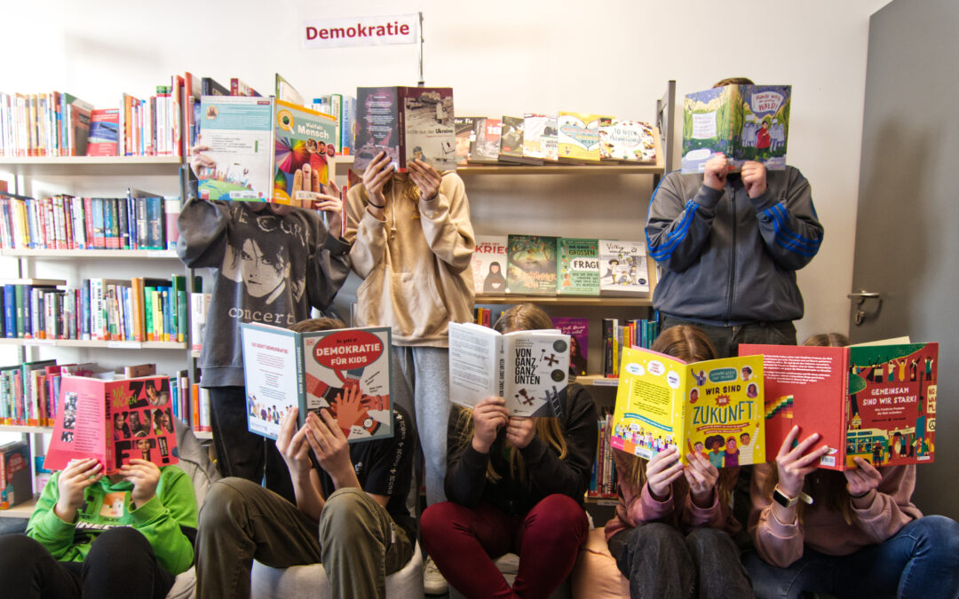Schülerinnen und Schüler vor den erworbenen Medien. Aus Datenschutzgründen halten sie Bücher vor ihr Gesicht.
